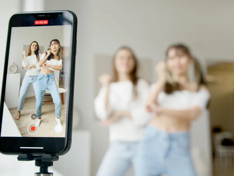 Two young women dance in front of a smartphone recording their choreography.  Two other women watch in the background.